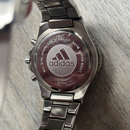 Adidas VS Chronograph 10-5010 Analog Watch is a silver-tone metal timepiece featuring a metallic blue dial with day and seconds indicators. The watch includes three sub-dials, a detailed seconds track, and three side buttons for chronograph functions. Designed with a sturdy, thick case, it comes with an extra link for adjustable sizing.