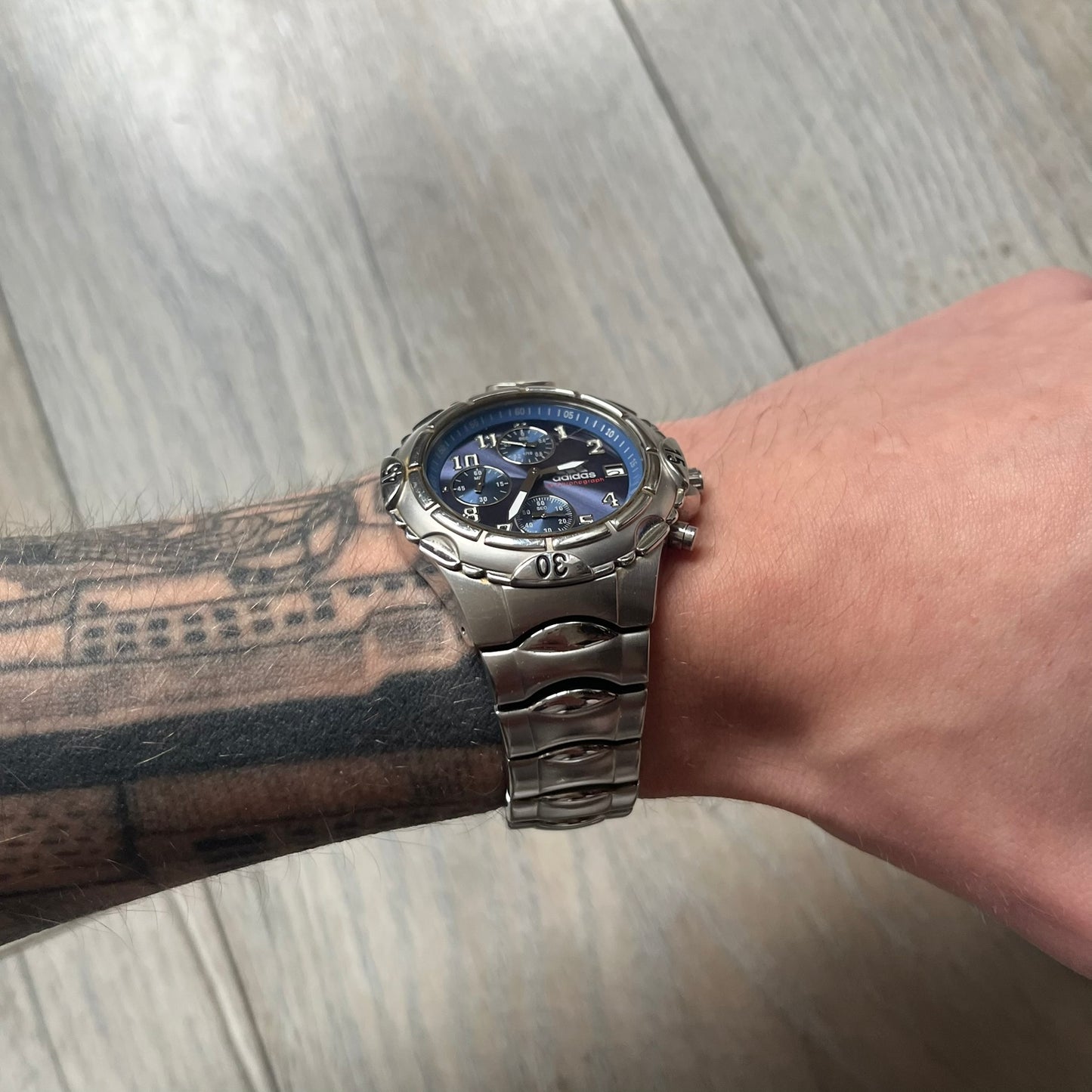 Adidas VS Chronograph 10-5010 Analog Watch is a silver-tone metal timepiece featuring a metallic blue dial with day and seconds indicators. The watch includes three sub-dials, a detailed seconds track, and three side buttons for chronograph functions. Designed with a sturdy, thick case, it comes with an extra link for adjustable sizing.