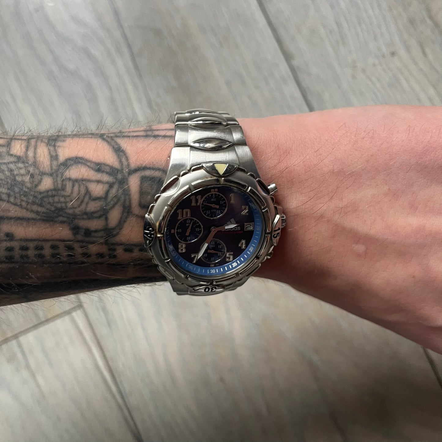 Adidas VS Chronograph 10-5010 Analog Watch is a silver-tone metal timepiece featuring a metallic blue dial with day and seconds indicators. The watch includes three sub-dials, a detailed seconds track, and three side buttons for chronograph functions. Designed with a sturdy, thick case, it comes with an extra link for adjustable sizing.