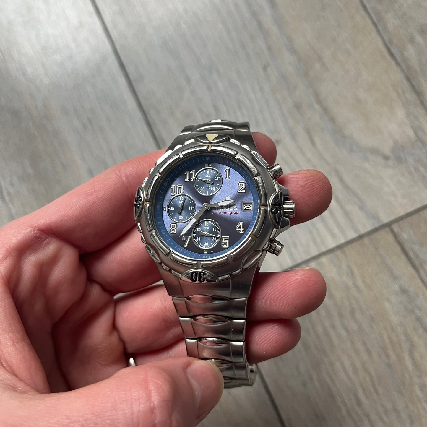 Adidas VS Chronograph 10-5010 Analog Watch is a silver-tone metal timepiece featuring a metallic blue dial with day and seconds indicators. The watch includes three sub-dials, a detailed seconds track, and three side buttons for chronograph functions. Designed with a sturdy, thick case, it comes with an extra link for adjustable sizing.