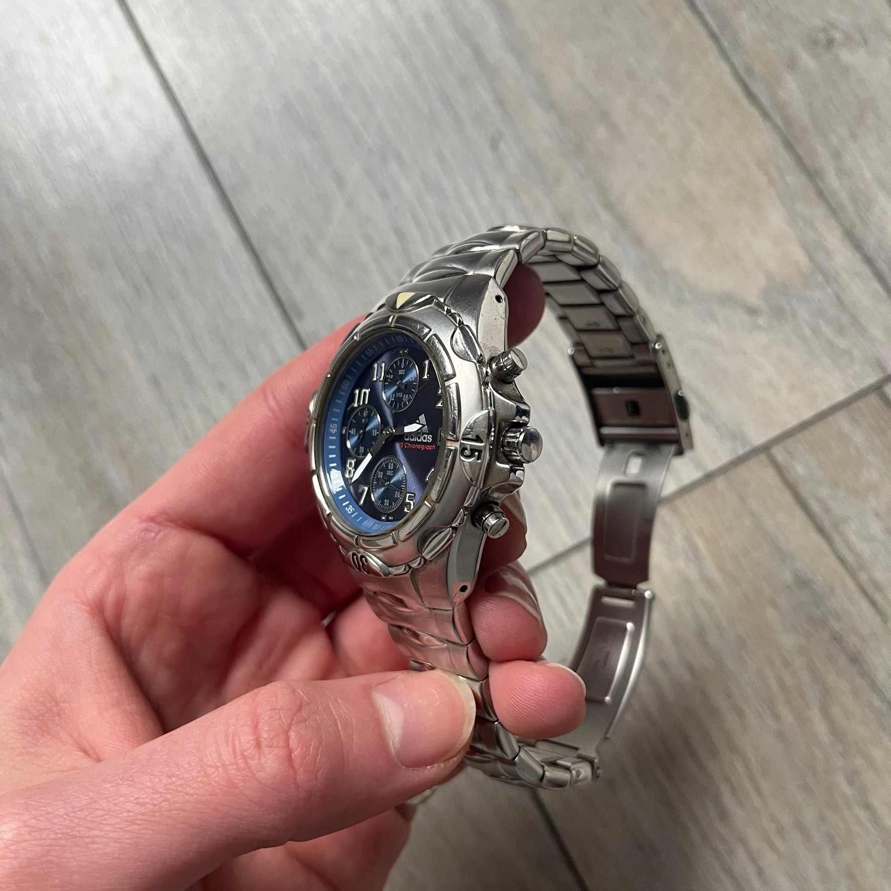 Adidas VS Chronograph 10-5010 Analog Watch is a silver-tone metal timepiece featuring a metallic blue dial with day and seconds indicators. The watch includes three sub-dials, a detailed seconds track, and three side buttons for chronograph functions. Designed with a sturdy, thick case, it comes with an extra link for adjustable sizing.