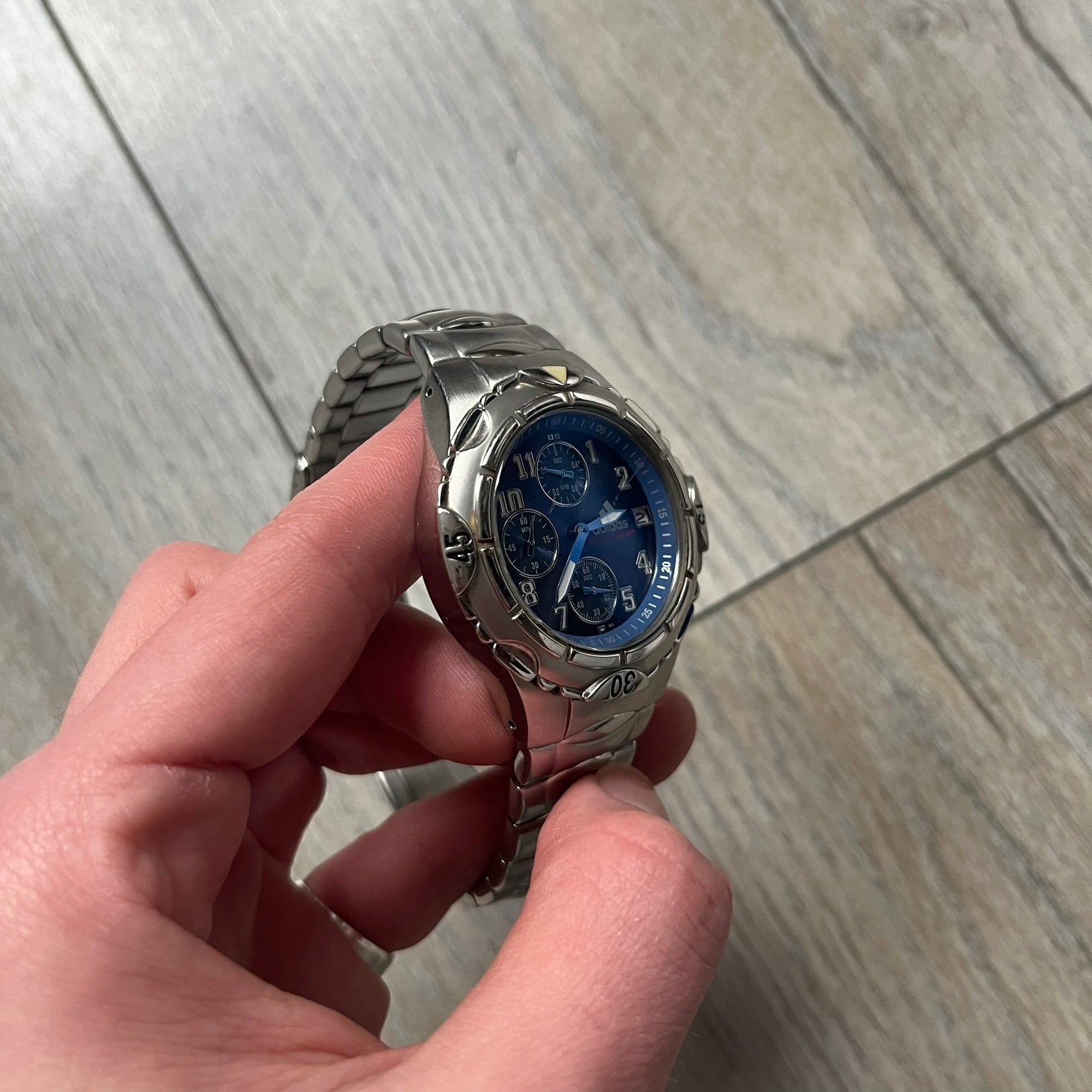 Adidas VS Chronograph 10-5010 Analog Watch is a silver-tone metal timepiece featuring a metallic blue dial with day and seconds indicators. The watch includes three sub-dials, a detailed seconds track, and three side buttons for chronograph functions. Designed with a sturdy, thick case, it comes with an extra link for adjustable sizing.