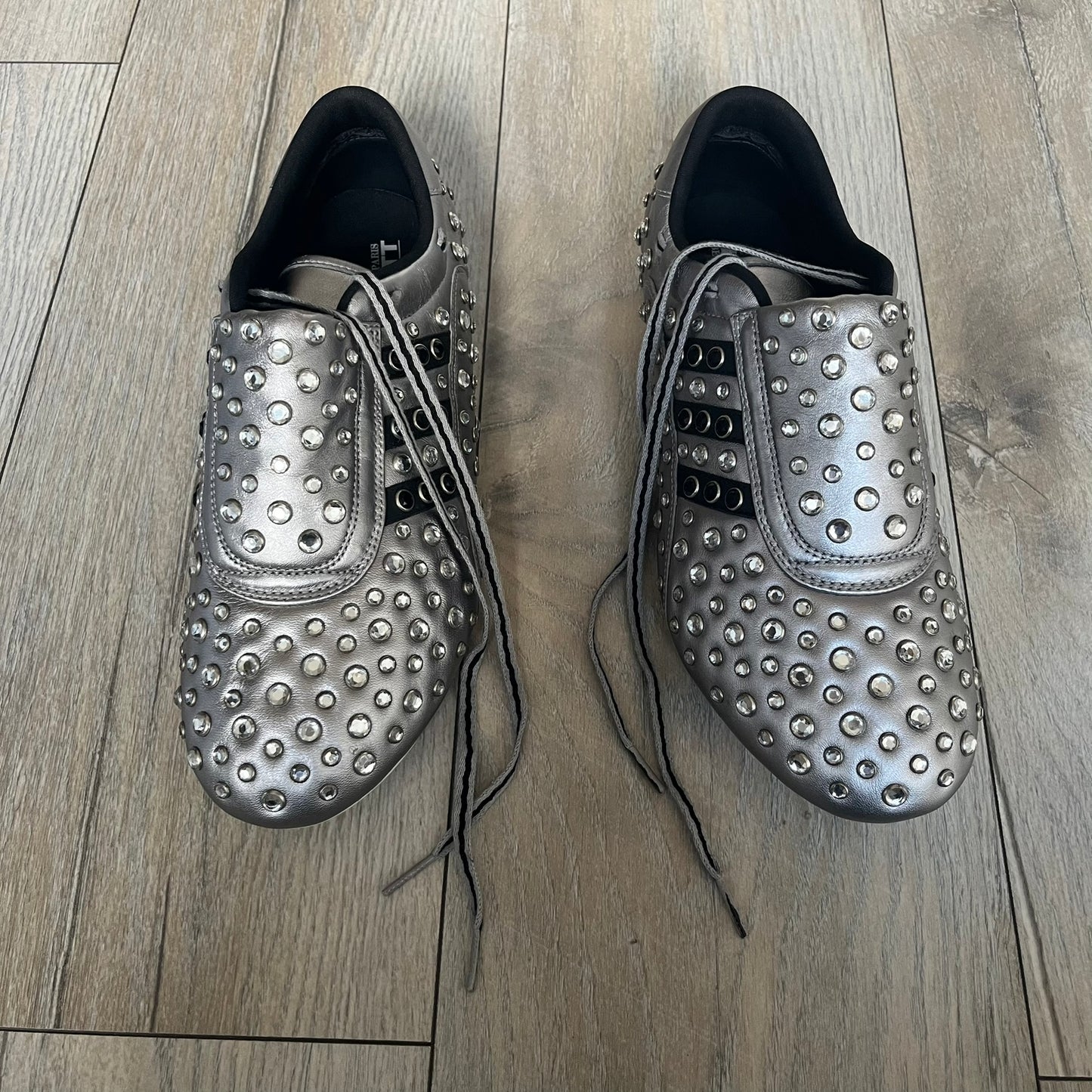 Adidas x Jeremy Scott 2010 Sample “Rockstar” Shoes (Article G16255) are a rare sample pair from the Spring/Summer 2010 collection, produced in June 2009. The shoes feature a metallic leather upper fully covered in circular rhinestone studs, with black Adidas three-stripe detailing on the sides and white soles. This model is built on a slim taekwondo-style silhouette and includes a yellow sample tag card.