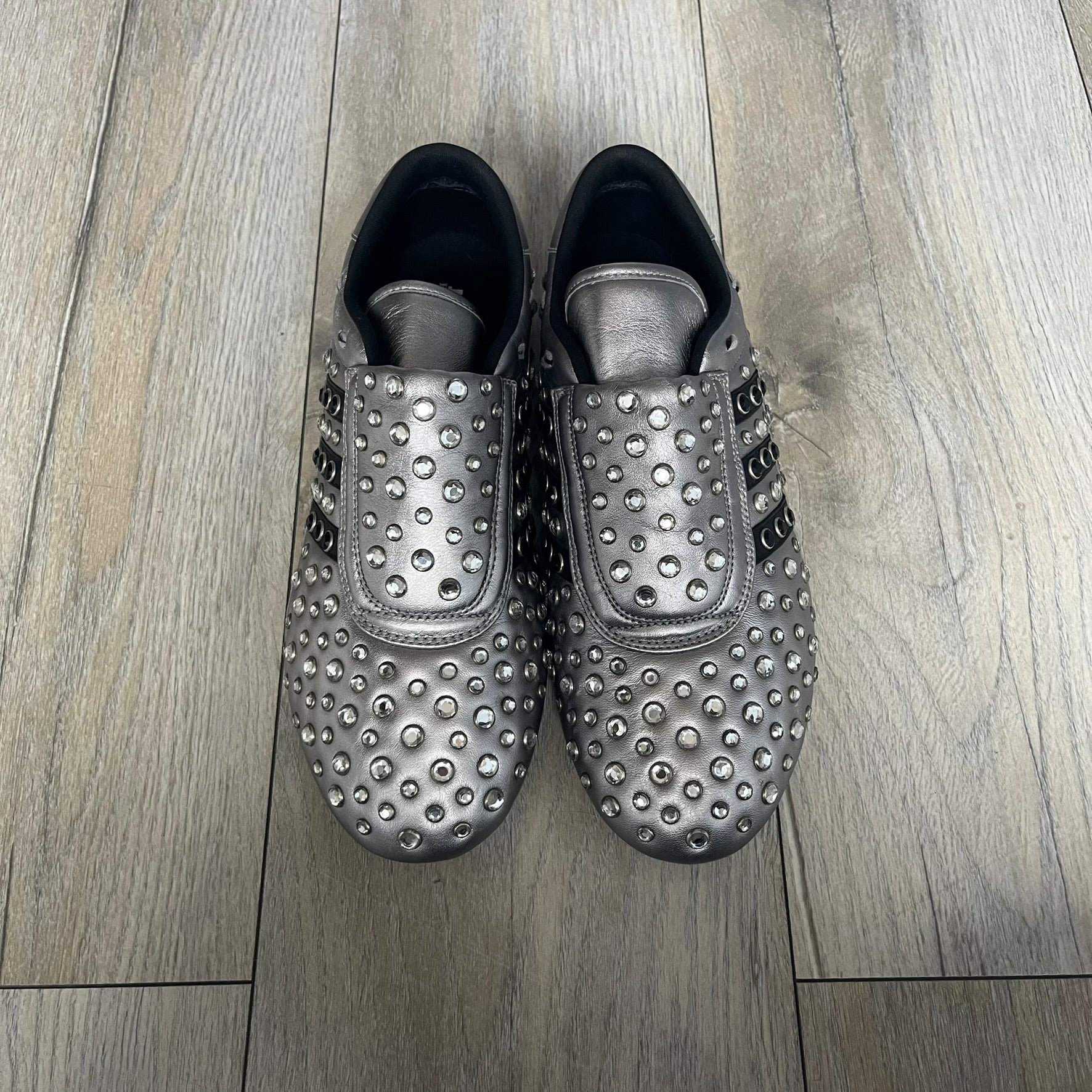 Adidas x Jeremy Scott 2010 Sample “Rockstar” Shoes (Article G16255) are a rare sample pair from the Spring/Summer 2010 collection, produced in June 2009. The shoes feature a metallic leather upper fully covered in circular rhinestone studs, with black Adidas three-stripe detailing on the sides and white soles. This model is built on a slim taekwondo-style silhouette and includes a yellow sample tag card.