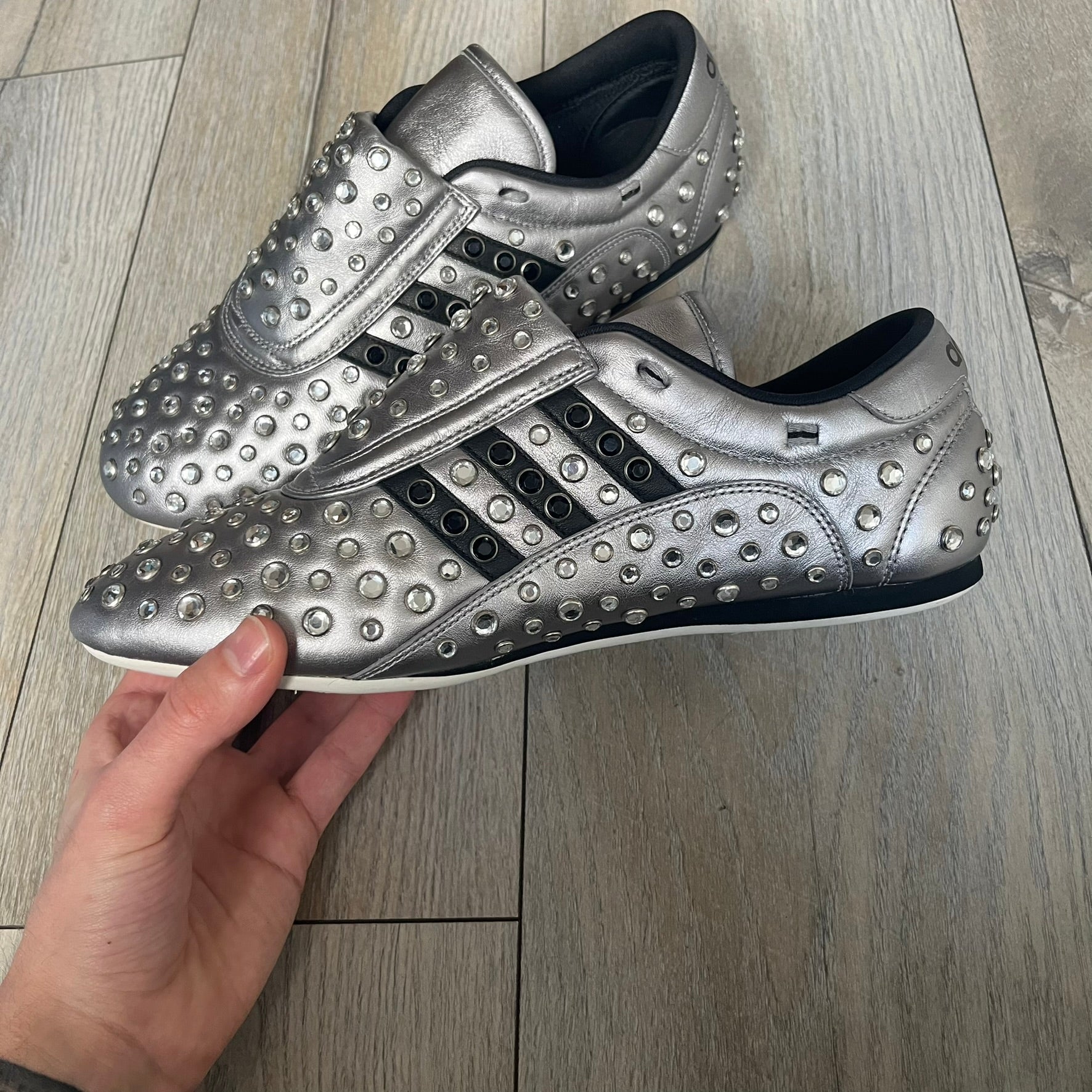 Adidas x Jeremy Scott 2010 Sample “Rockstar” Shoes (Article G16255) are a rare sample pair from the Spring/Summer 2010 collection, produced in June 2009. The shoes feature a metallic leather upper fully covered in circular rhinestone studs, with black Adidas three-stripe detailing on the sides and white soles. This model is built on a slim taekwondo-style silhouette and includes a yellow sample tag card.