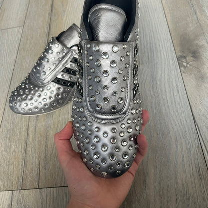 Adidas x Jeremy Scott 2010 Sample “Rockstar” Shoes (Article G16255) are a rare sample pair from the Spring/Summer 2010 collection, produced in June 2009. The shoes feature a metallic leather upper fully covered in circular rhinestone studs, with black Adidas three-stripe detailing on the sides and white soles. This model is built on a slim taekwondo-style silhouette and includes a yellow sample tag card.