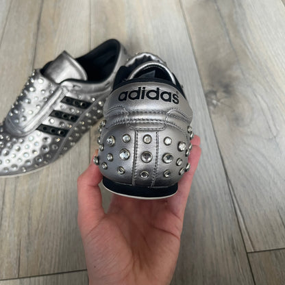 Adidas x Jeremy Scott 2010 Sample “Rockstar” Shoes (Article G16255) are a rare sample pair from the Spring/Summer 2010 collection, produced in June 2009. The shoes feature a metallic leather upper fully covered in circular rhinestone studs, with black Adidas three-stripe detailing on the sides and white soles. This model is built on a slim taekwondo-style silhouette and includes a yellow sample tag card.