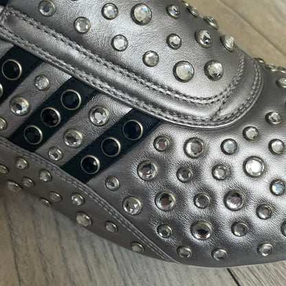 Adidas x Jeremy Scott 2010 Sample “Rockstar” Shoes (Article G16255) are a rare sample pair from the Spring/Summer 2010 collection, produced in June 2009. The shoes feature a metallic leather upper fully covered in circular rhinestone studs, with black Adidas three-stripe detailing on the sides and white soles. This model is built on a slim taekwondo-style silhouette and includes a yellow sample tag card.