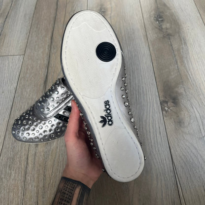 Adidas x Jeremy Scott 2010 Sample “Rockstar” Shoes (Article G16255) are a rare sample pair from the Spring/Summer 2010 collection, produced in June 2009. The shoes feature a metallic leather upper fully covered in circular rhinestone studs, with black Adidas three-stripe detailing on the sides and white soles. This model is built on a slim taekwondo-style silhouette and includes a yellow sample tag card.