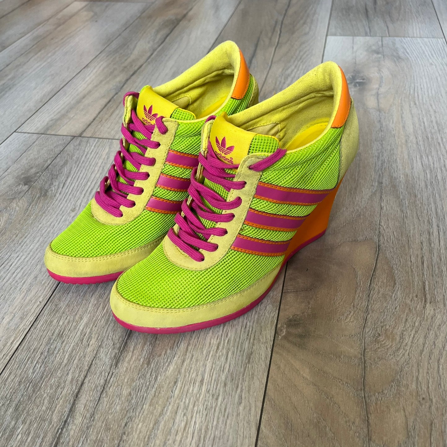 Adidas x Jeremy Scott 2013 Arrow Wedge Neoncamo Platform Shoes (G96747) are a high-platform sneaker built on a track spike-inspired silhouette. The upper features a neon camo color scheme combining green, purple, and orange tones, with light green suede panels on the toe and heel. The shoe includes purple laces, a purple outsole, and a tall orange wedge-style platform at the heel with a textured, waffled sole construction.