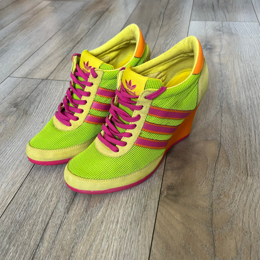 Adidas x Jeremy Scott 2013 Arrow Wedge Neoncamo Platform Shoes (G96747) are a high-platform sneaker built on a track spike-inspired silhouette. The upper features a neon camo color scheme combining green, purple, and orange tones, with light green suede panels on the toe and heel. The shoe includes purple laces, a purple outsole, and a tall orange wedge-style platform at the heel with a textured, waffled sole construction.