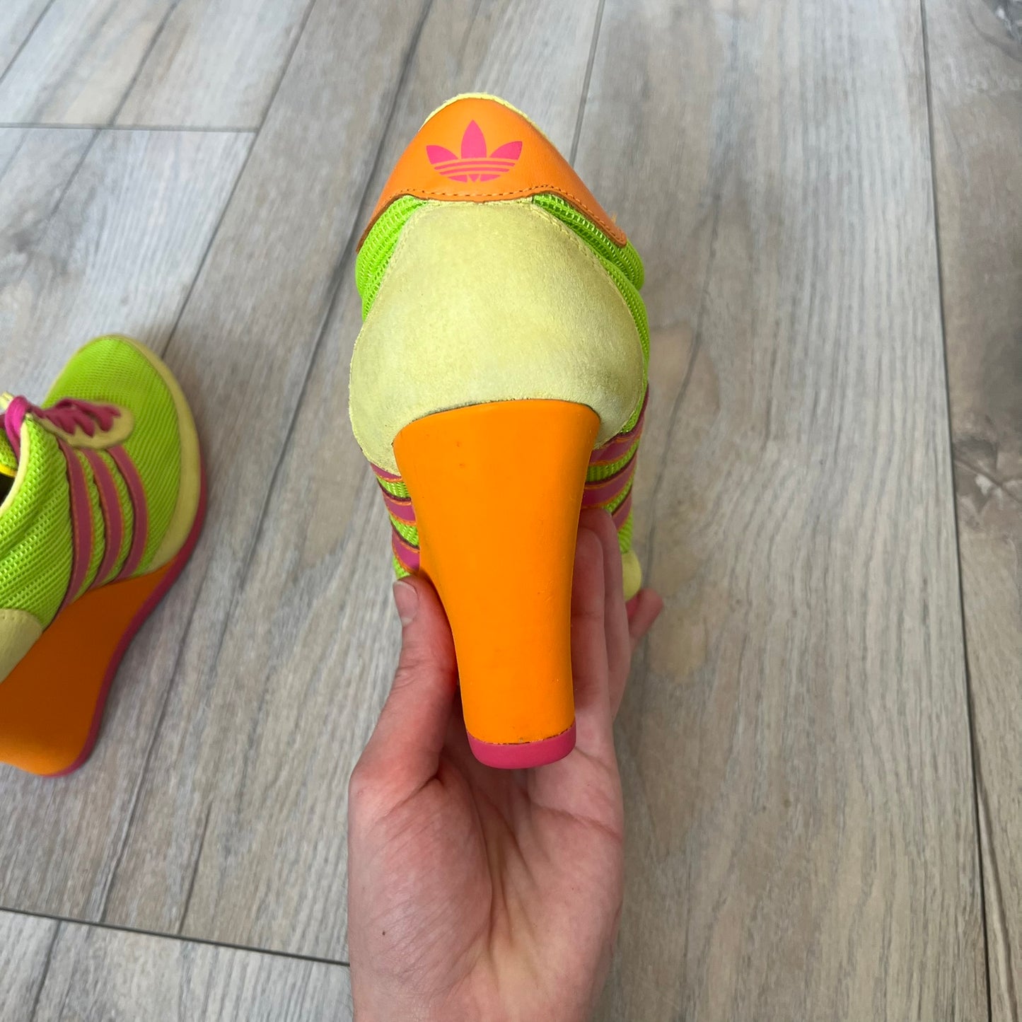 Adidas x Jeremy Scott 2013 Arrow Wedge Neoncamo Platform Shoes (G96747) are a high-platform sneaker built on a track spike-inspired silhouette. The upper features a neon camo color scheme combining green, purple, and orange tones, with light green suede panels on the toe and heel. The shoe includes purple laces, a purple outsole, and a tall orange wedge-style platform at the heel with a textured, waffled sole construction.