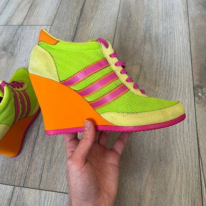 Adidas x Jeremy Scott 2013 Arrow Wedge Neoncamo Platform Shoes (G96747) are a high-platform sneaker built on a track spike-inspired silhouette. The upper features a neon camo color scheme combining green, purple, and orange tones, with light green suede panels on the toe and heel. The shoe includes purple laces, a purple outsole, and a tall orange wedge-style platform at the heel with a textured, waffled sole construction.