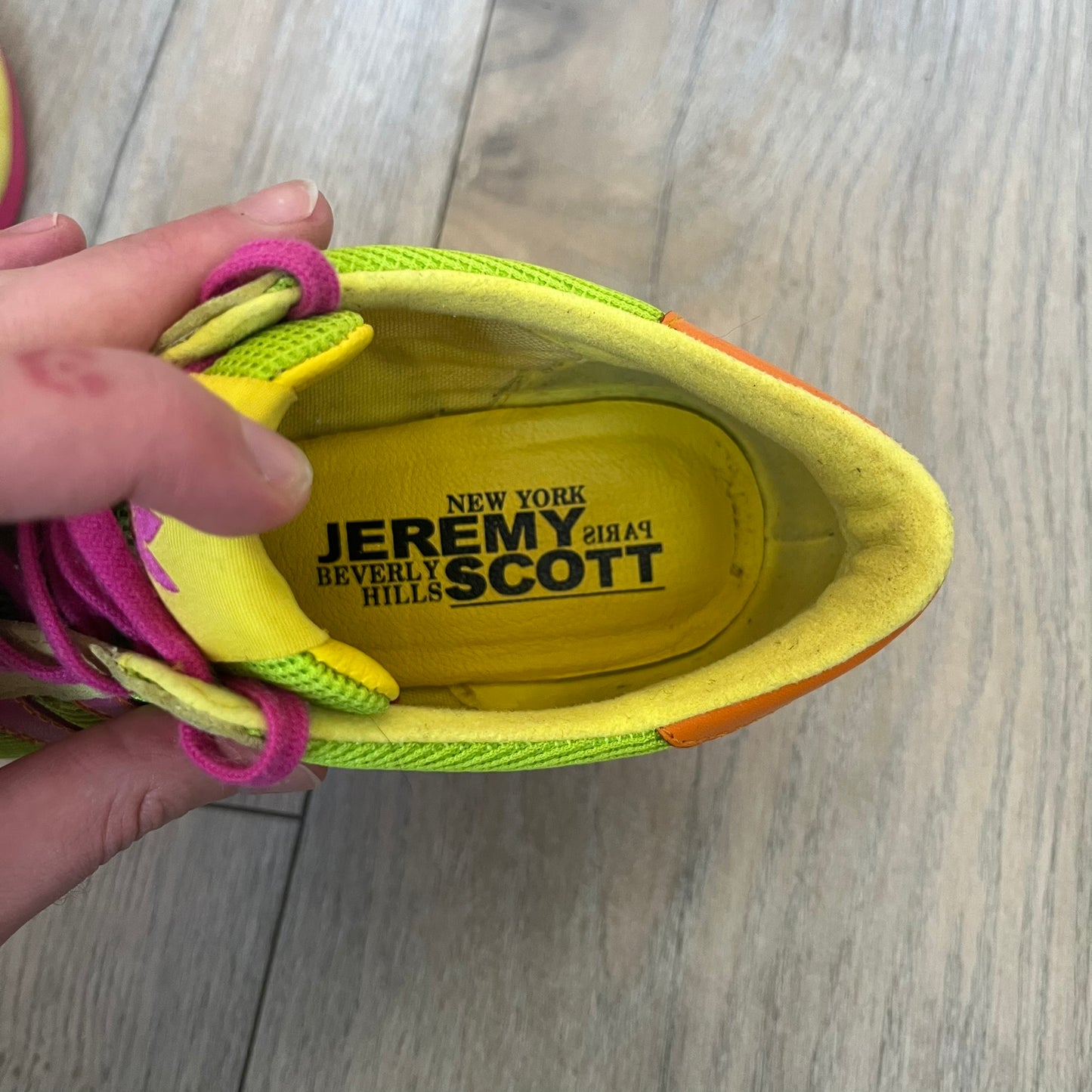 Adidas x Jeremy Scott 2013 Arrow Wedge Neoncamo Platform Shoes (G96747) are a high-platform sneaker built on a track spike-inspired silhouette. The upper features a neon camo color scheme combining green, purple, and orange tones, with light green suede panels on the toe and heel. The shoe includes purple laces, a purple outsole, and a tall orange wedge-style platform at the heel with a textured, waffled sole construction.