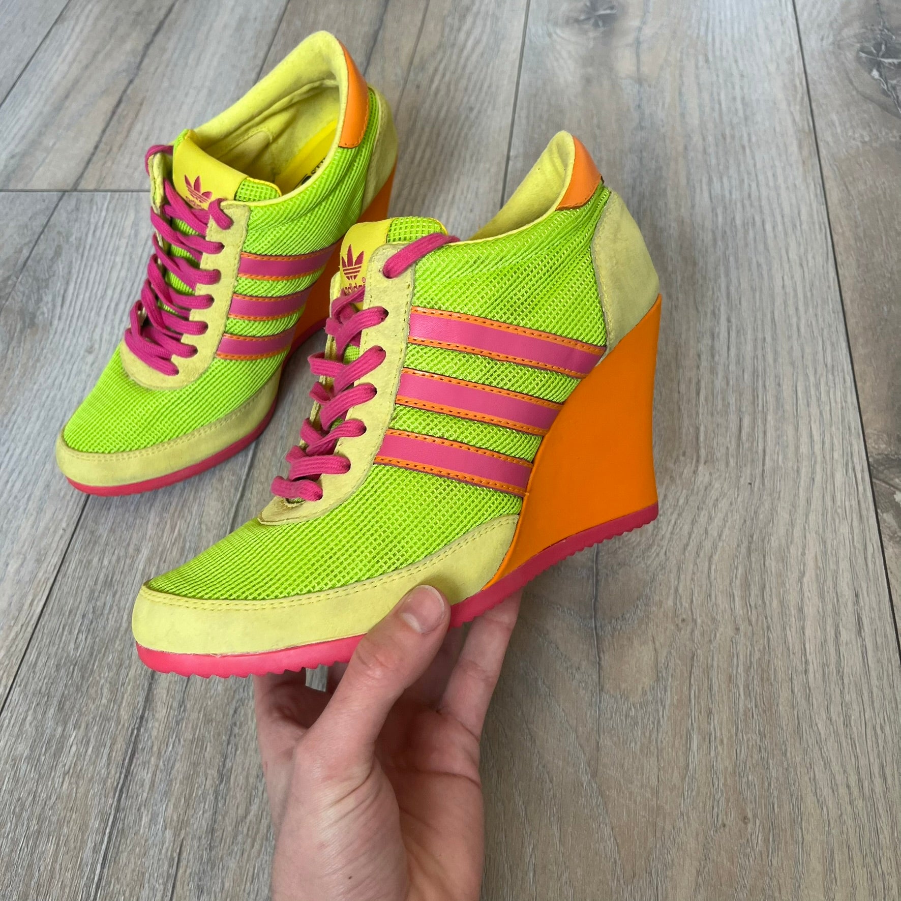 Adidas x Jeremy Scott 2013 Arrow Wedge Neoncamo Platform Shoes (G96747) are a high-platform sneaker built on a track spike-inspired silhouette. The upper features a neon camo color scheme combining green, purple, and orange tones, with light green suede panels on the toe and heel. The shoe includes purple laces, a purple outsole, and a tall orange wedge-style platform at the heel with a textured, waffled sole construction.
