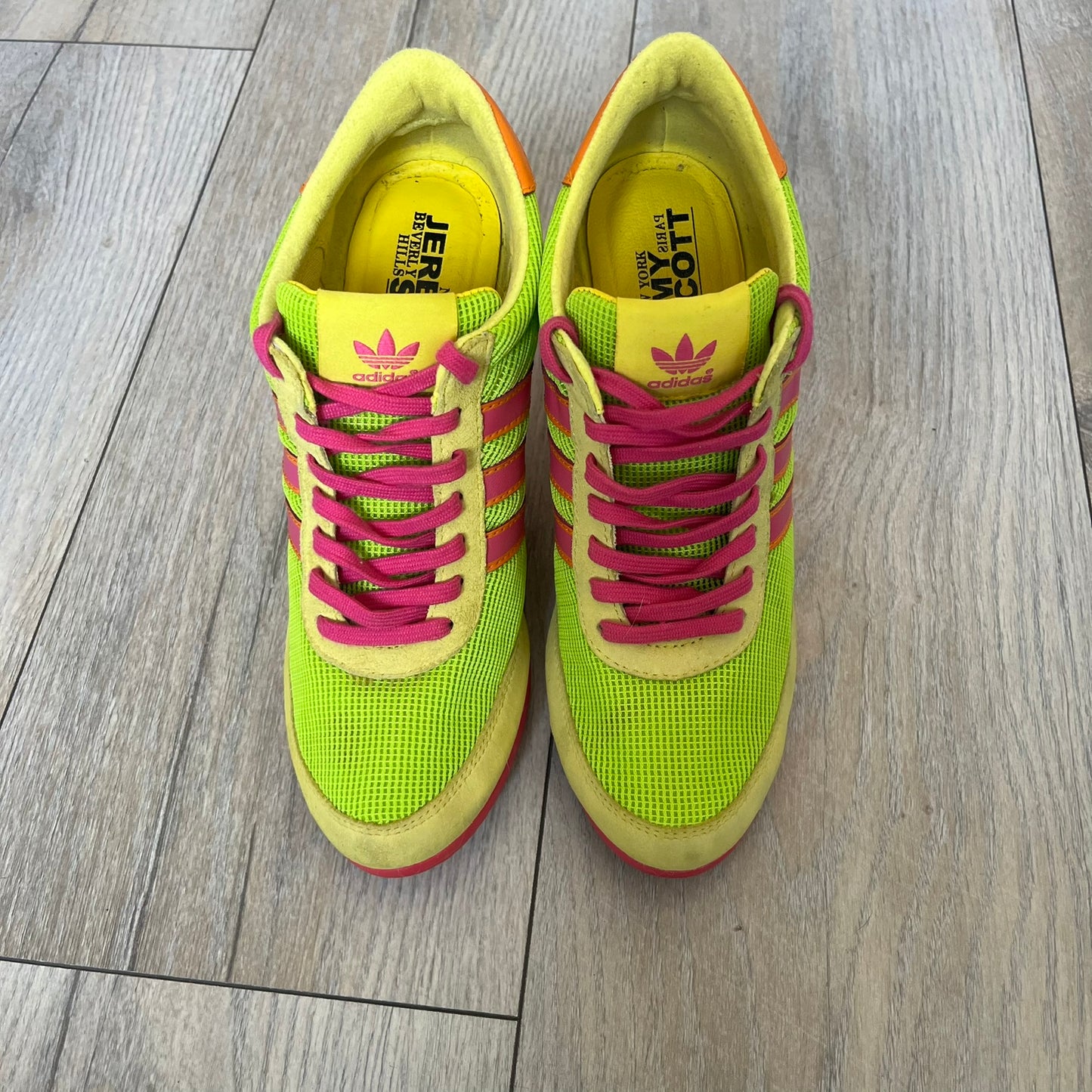 Adidas x Jeremy Scott 2013 Arrow Wedge Neoncamo Platform Shoes (G96747) are a high-platform sneaker built on a track spike-inspired silhouette. The upper features a neon camo color scheme combining green, purple, and orange tones, with light green suede panels on the toe and heel. The shoe includes purple laces, a purple outsole, and a tall orange wedge-style platform at the heel with a textured, waffled sole construction.