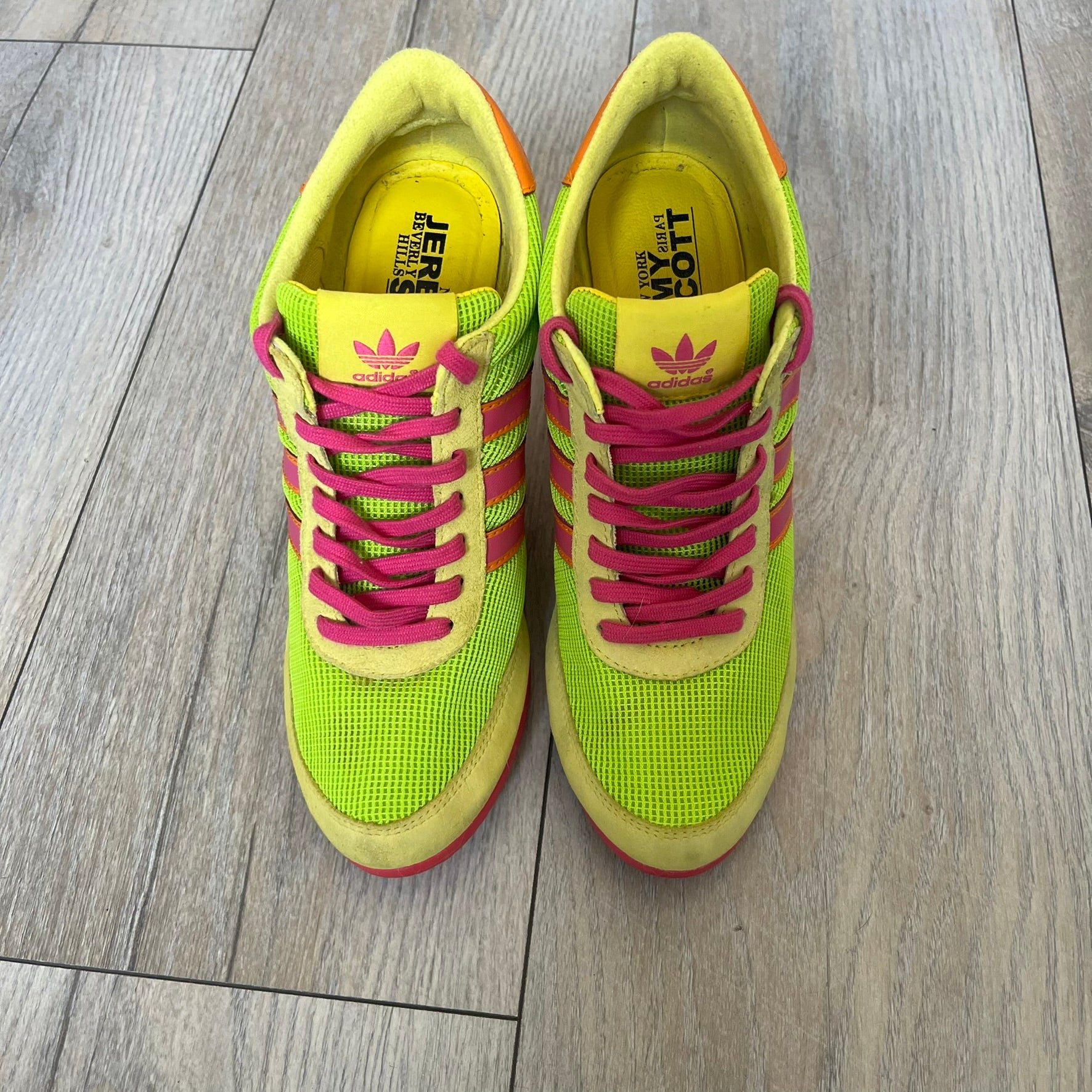 Adidas x Jeremy Scott 2013 Arrow Wedge Neoncamo Platform Shoes (G96747) are a high-platform sneaker built on a track spike-inspired silhouette. The upper features a neon camo color scheme combining green, purple, and orange tones, with light green suede panels on the toe and heel. The shoe includes purple laces, a purple outsole, and a tall orange wedge-style platform at the heel with a textured, waffled sole construction.