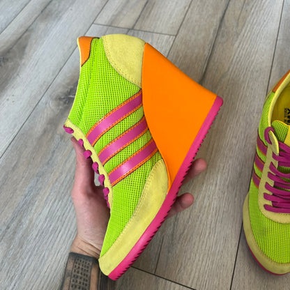 Adidas x Jeremy Scott 2013 Arrow Wedge Neoncamo Platform Shoes (G96747) are a high-platform sneaker built on a track spike-inspired silhouette. The upper features a neon camo color scheme combining green, purple, and orange tones, with light green suede panels on the toe and heel. The shoe includes purple laces, a purple outsole, and a tall orange wedge-style platform at the heel with a textured, waffled sole construction.