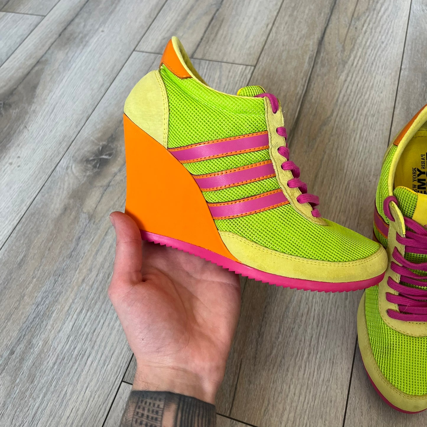 Adidas x Jeremy Scott 2013 Arrow Wedge Neoncamo Platform Shoes (G96747) are a high-platform sneaker built on a track spike-inspired silhouette. The upper features a neon camo color scheme combining green, purple, and orange tones, with light green suede panels on the toe and heel. The shoe includes purple laces, a purple outsole, and a tall orange wedge-style platform at the heel with a textured, waffled sole construction.