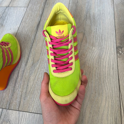Adidas x Jeremy Scott 2013 Arrow Wedge Neoncamo Platform Shoes (G96747) are a high-platform sneaker built on a track spike-inspired silhouette. The upper features a neon camo color scheme combining green, purple, and orange tones, with light green suede panels on the toe and heel. The shoe includes purple laces, a purple outsole, and a tall orange wedge-style platform at the heel with a textured, waffled sole construction.