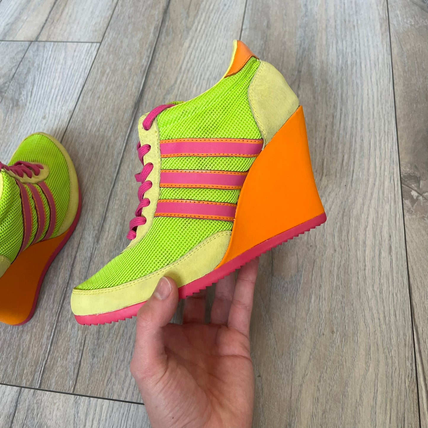 Adidas x Jeremy Scott 2013 Arrow Wedge Neoncamo Platform Shoes (G96747) are a high-platform sneaker built on a track spike-inspired silhouette. The upper features a neon camo color scheme combining green, purple, and orange tones, with light green suede panels on the toe and heel. The shoe includes purple laces, a purple outsole, and a tall orange wedge-style platform at the heel with a textured, waffled sole construction.
