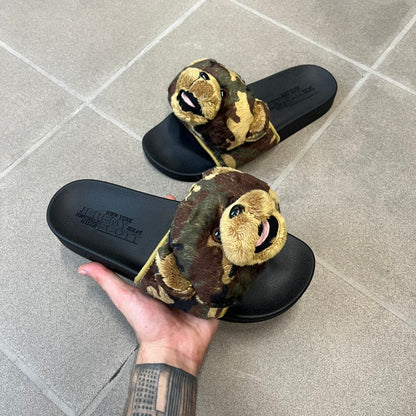 Adidas x Jeremy Scott Adilette Teddy Bear Slides (H02882) were released on November 11, 2021. They feature a black slip-on Adilette silhouette with a camouflage faux fur bandage strap topped by a plush teddy bear head. The strap is lined with leather, while the molded footbed and EVA midsole provide cushioning. Jeremy Scott branding is debossed on the footbed, completing the collaboration design.
