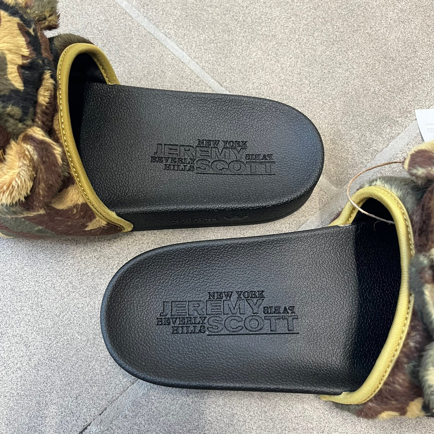 Adidas x Jeremy Scott Adilette Teddy Bear Slides (H02882) were released on November 11, 2021. They feature a black slip-on Adilette silhouette with a camouflage faux fur bandage strap topped by a plush teddy bear head. The strap is lined with leather, while the molded footbed and EVA midsole provide cushioning. Jeremy Scott branding is debossed on the footbed, completing the collaboration design.