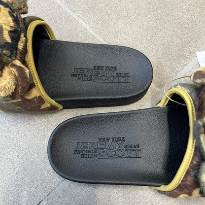 Adidas x Jeremy Scott Adilette Teddy Bear Slides (H02882) were released on November 11, 2021. They feature a black slip-on Adilette silhouette with a camouflage faux fur bandage strap topped by a plush teddy bear head. The strap is lined with leather, while the molded footbed and EVA midsole provide cushioning. Jeremy Scott branding is debossed on the footbed, completing the collaboration design.