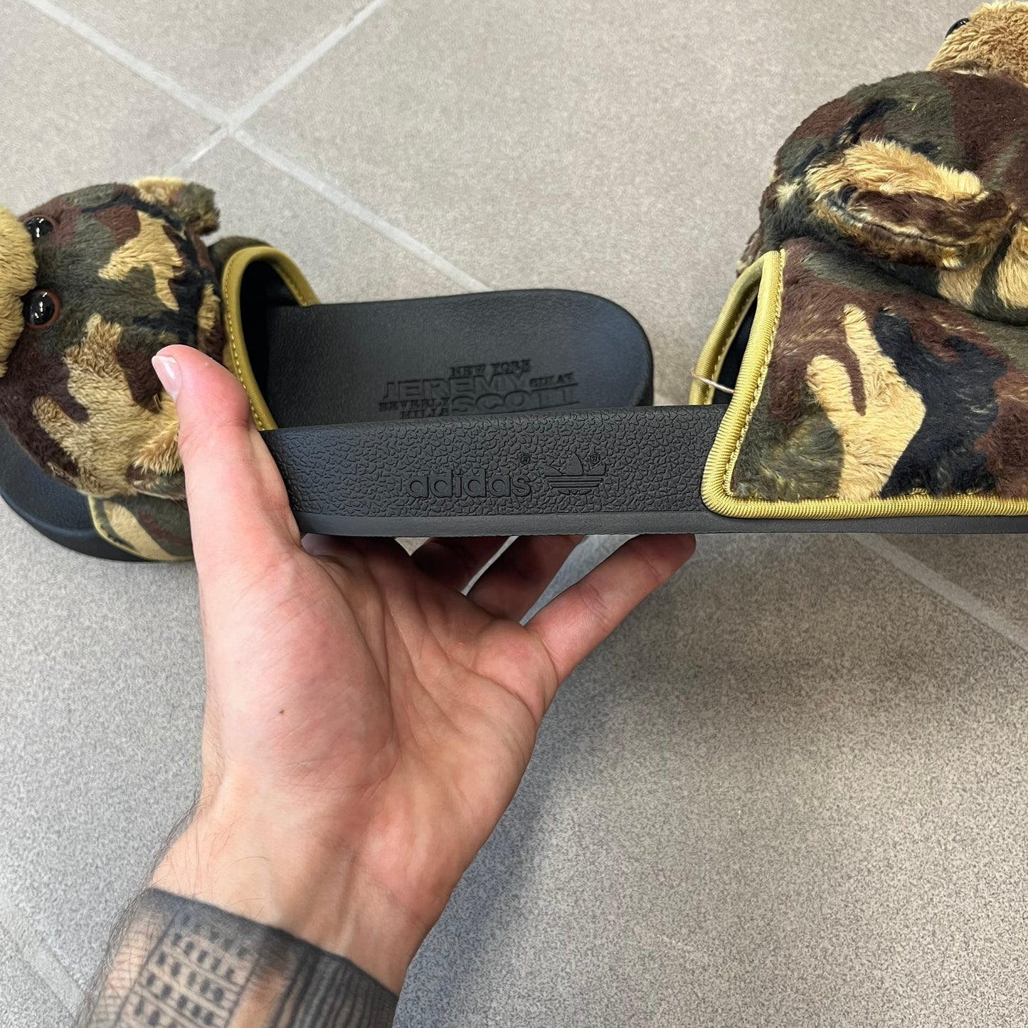 Adidas x Jeremy Scott Adilette Teddy Bear Slides (H02882) were released on November 11, 2021. They feature a black slip-on Adilette silhouette with a camouflage faux fur bandage strap topped by a plush teddy bear head. The strap is lined with leather, while the molded footbed and EVA midsole provide cushioning. Jeremy Scott branding is debossed on the footbed, completing the collaboration design.