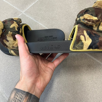 Adidas x Jeremy Scott Adilette Teddy Bear Slides (H02882) were released on November 11, 2021. They feature a black slip-on Adilette silhouette with a camouflage faux fur bandage strap topped by a plush teddy bear head. The strap is lined with leather, while the molded footbed and EVA midsole provide cushioning. Jeremy Scott branding is debossed on the footbed, completing the collaboration design.