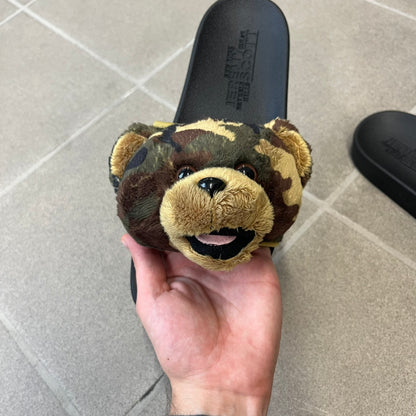 Adidas x Jeremy Scott Adilette Teddy Bear Slides (H02882) were released on November 11, 2021. They feature a black slip-on Adilette silhouette with a camouflage faux fur bandage strap topped by a plush teddy bear head. The strap is lined with leather, while the molded footbed and EVA midsole provide cushioning. Jeremy Scott branding is debossed on the footbed, completing the collaboration design.