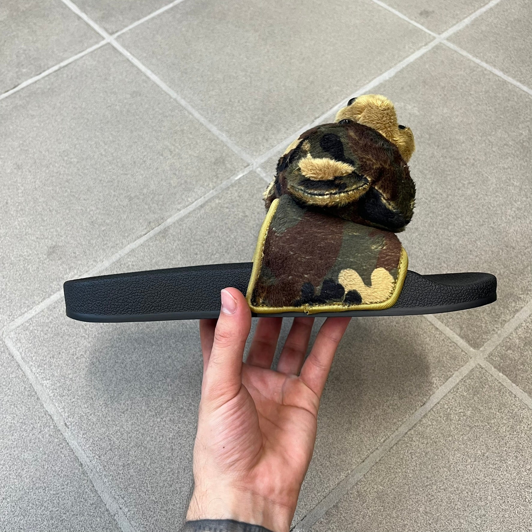 Adidas x Jeremy Scott Adilette Teddy Bear Slides (H02882) were released on November 11, 2021. They feature a black slip-on Adilette silhouette with a camouflage faux fur bandage strap topped by a plush teddy bear head. The strap is lined with leather, while the molded footbed and EVA midsole provide cushioning. Jeremy Scott branding is debossed on the footbed, completing the collaboration design.