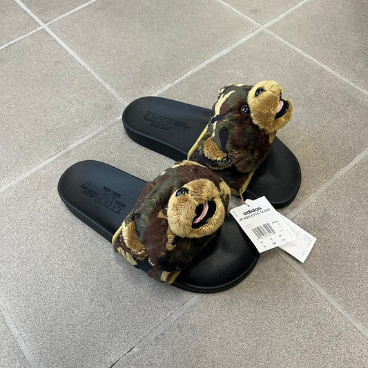 Adidas x Jeremy Scott Adilette Teddy Bear Slides (H02882) were released on November 11, 2021. They feature a black slip-on Adilette silhouette with a camouflage faux fur bandage strap topped by a plush teddy bear head. The strap is lined with leather, while the molded footbed and EVA midsole provide cushioning. Jeremy Scott branding is debossed on the footbed, completing the collaboration design.