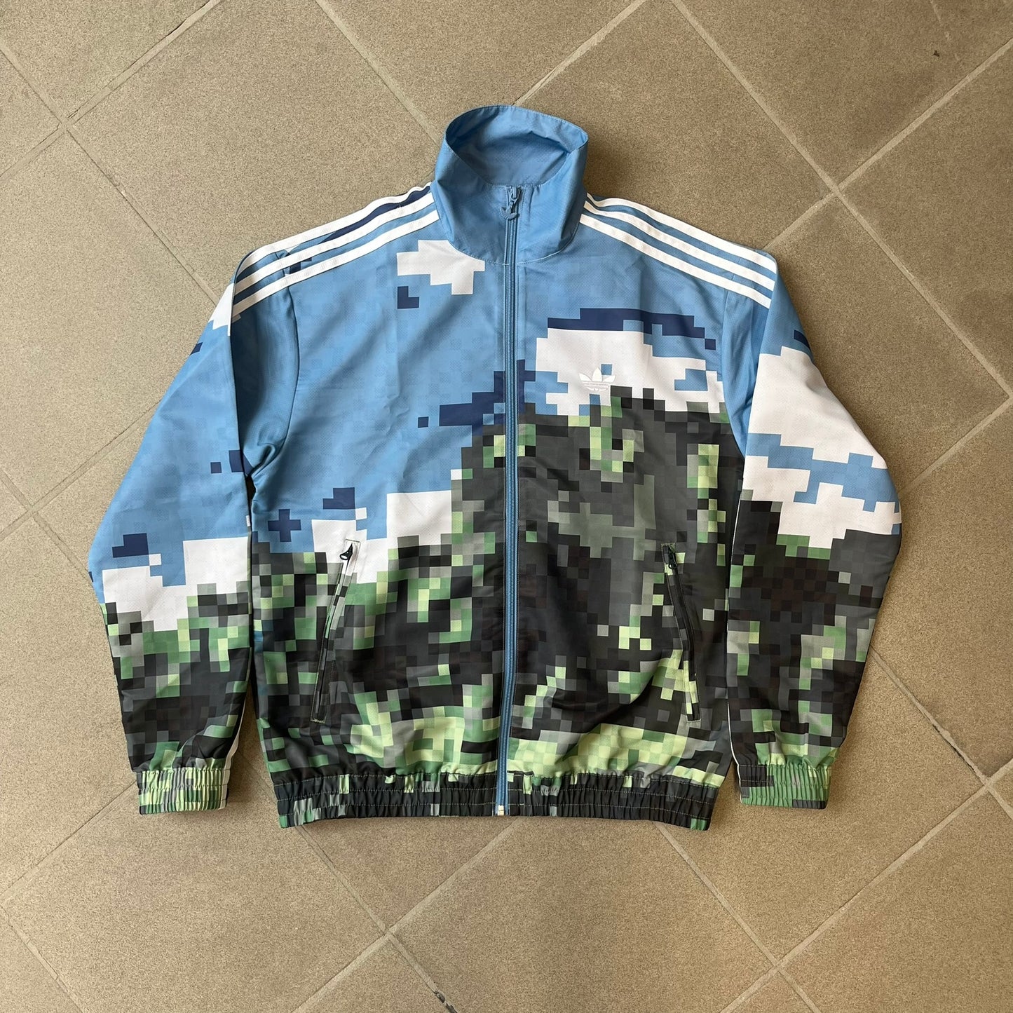 Adidas x Minecraft Movie SS25 Track Top (Style: JZ9921) is a Spring/Summer 2025 collaboration featuring an all-over Minecraft landscape print with a pixelated sky across the upper half and grass terrain across the lower half. The track jacket has white Three Stripes along the shoulders and sleeves, a full-zip front with a pixelated Trefoil logo zipper pull, and ribbed cuffs and hem. Finished in Hazy Blue, Magic Lime, and Tribe Green.
