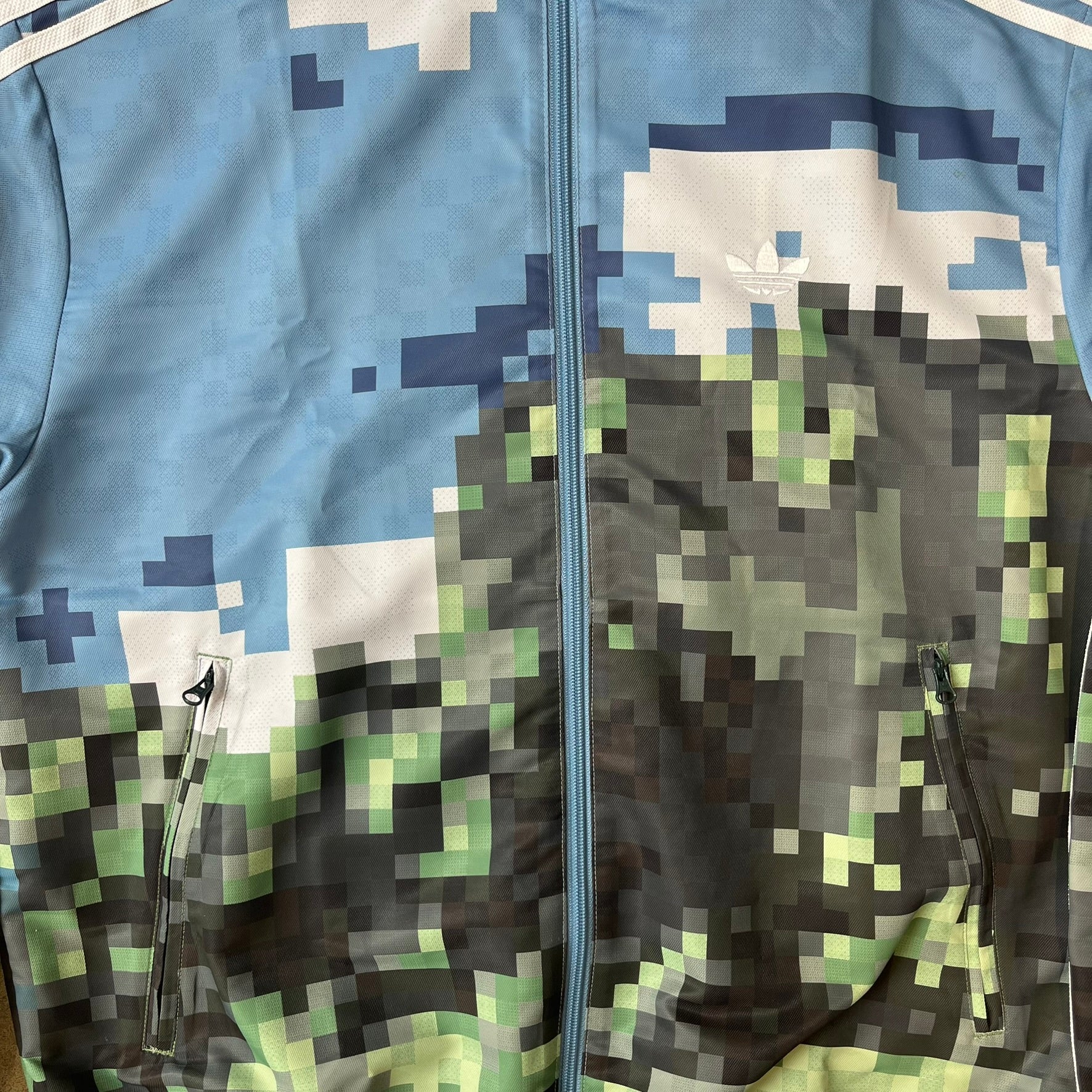 Adidas x Minecraft Movie SS25 Track Top (Style: JZ9921) is a Spring/Summer 2025 collaboration featuring an all-over Minecraft landscape print with a pixelated sky across the upper half and grass terrain across the lower half. The track jacket has white Three Stripes along the shoulders and sleeves, a full-zip front with a pixelated Trefoil logo zipper pull, and ribbed cuffs and hem. Finished in Hazy Blue, Magic Lime, and Tribe Green.