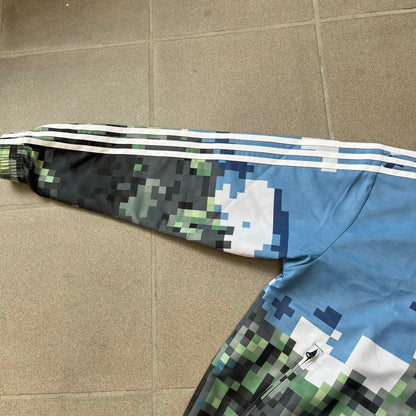 Adidas x Minecraft Movie SS25 Track Top (Style: JZ9921) is a Spring/Summer 2025 collaboration featuring an all-over Minecraft landscape print with a pixelated sky across the upper half and grass terrain across the lower half. The track jacket has white Three Stripes along the shoulders and sleeves, a full-zip front with a pixelated Trefoil logo zipper pull, and ribbed cuffs and hem. Finished in Hazy Blue, Magic Lime, and Tribe Green.