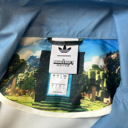 Adidas x Minecraft Movie SS25 Track Top (Style: JZ9921) is a Spring/Summer 2025 collaboration featuring an all-over Minecraft landscape print with a pixelated sky across the upper half and grass terrain across the lower half. The track jacket has white Three Stripes along the shoulders and sleeves, a full-zip front with a pixelated Trefoil logo zipper pull, and ribbed cuffs and hem. Finished in Hazy Blue, Magic Lime, and Tribe Green.