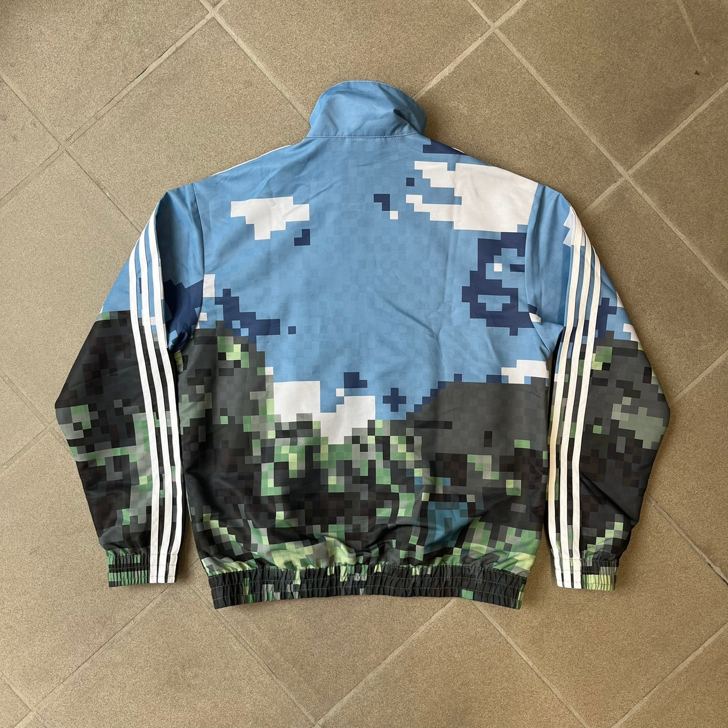 Adidas x Minecraft Movie SS25 Track Top (Style: JZ9921) is a Spring/Summer 2025 collaboration featuring an all-over Minecraft landscape print with a pixelated sky across the upper half and grass terrain across the lower half. The track jacket has white Three Stripes along the shoulders and sleeves, a full-zip front with a pixelated Trefoil logo zipper pull, and ribbed cuffs and hem. Finished in Hazy Blue, Magic Lime, and Tribe Green.