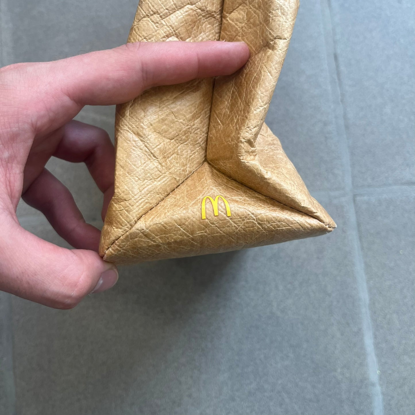 Alexander Wang x McDonald’s 2019 Golden Lunch Bag is a limited edition collaboration accessory made from thick waterproof DuPont Tyvek paper with a leather-like texture. Designed to resemble a classic McDonald’s takeaway bag, it features a small white Alexander Wang logo on the front and a subtle McDonald’s Golden Arches logo on the side. The piece combines fast-food nostalgia with Alexander Wang’s minimalist luxury approach.
