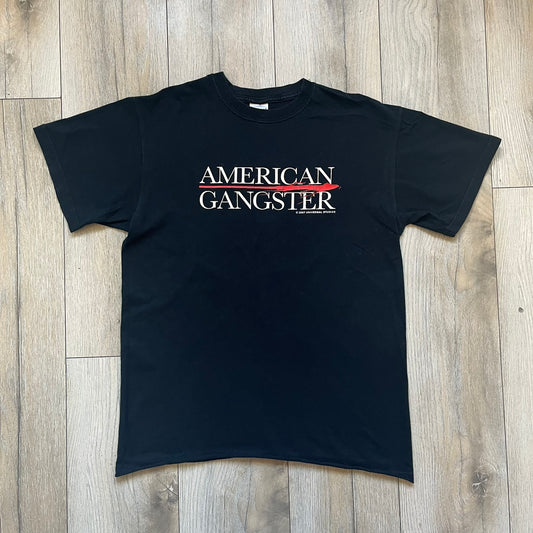 American Gangster 2007 Promo Tee is a black short-sleeve T-shirt made on a Gildan blank. The front features a white “American Gangster” logo with a red blood line between the words, and a small 2007 Universal Studios licensing print beneath. This shirt was created as official promotional merchandise for the 2007 film.