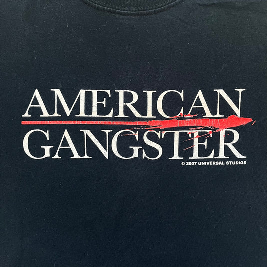 American Gangster 2007 Promo Tee is a black short-sleeve T-shirt made on a Gildan blank. The front features a white “American Gangster” logo with a red blood line between the words, and a small 2007 Universal Studios licensing print beneath. This shirt was created as official promotional merchandise for the 2007 film.