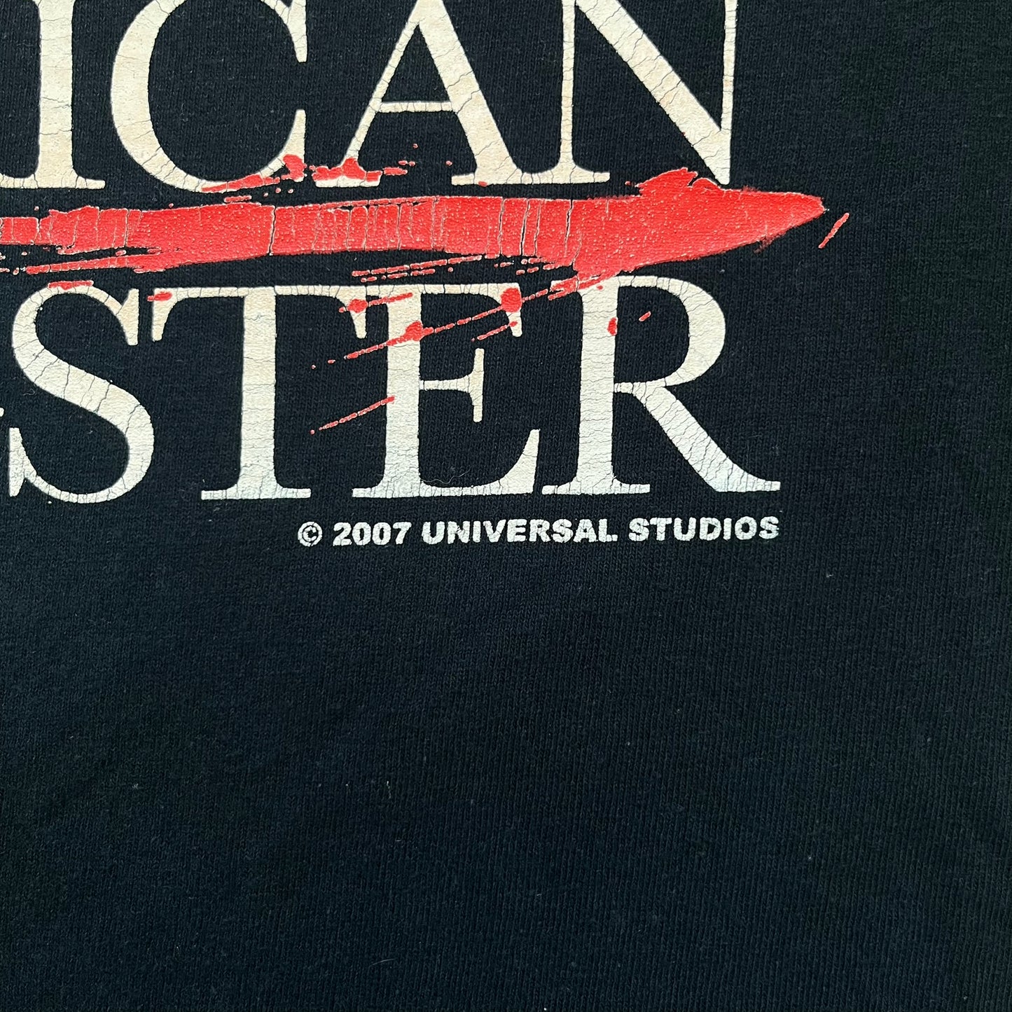 American Gangster 2007 Promo Tee is a black short-sleeve T-shirt made on a Gildan blank. The front features a white “American Gangster” logo with a red blood line between the words, and a small 2007 Universal Studios licensing print beneath. This shirt was created as official promotional merchandise for the 2007 film.