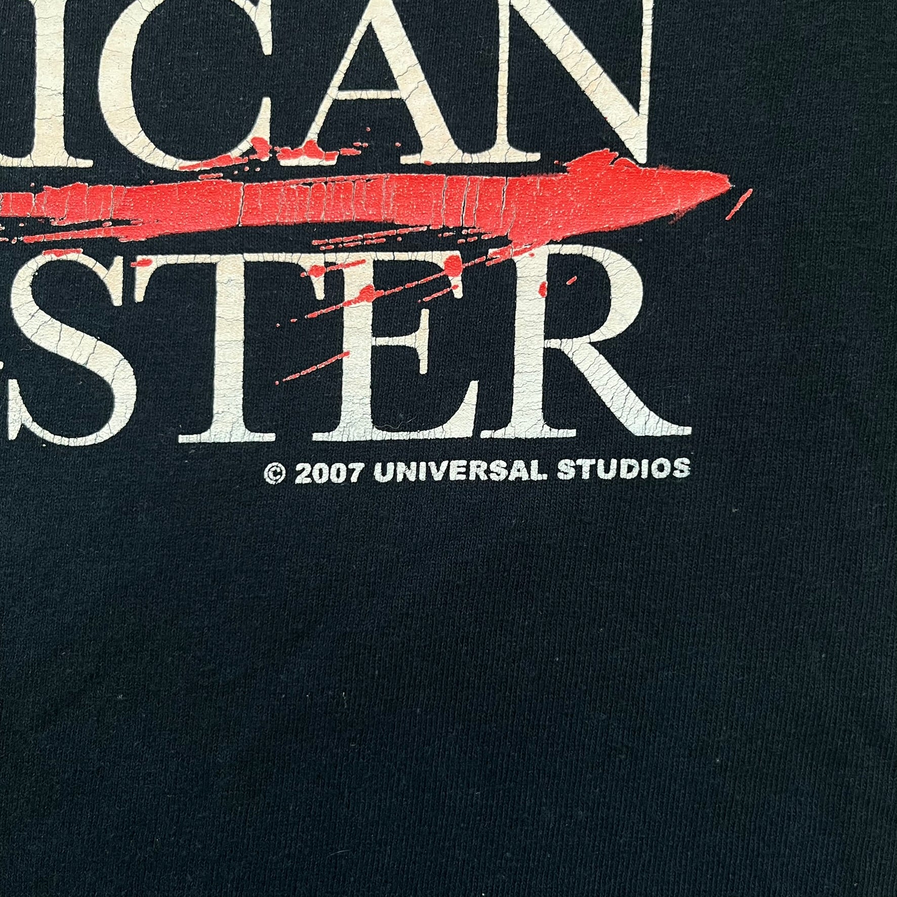 American Gangster 2007 Promo Tee is a black short-sleeve T-shirt made on a Gildan blank. The front features a white “American Gangster” logo with a red blood line between the words, and a small 2007 Universal Studios licensing print beneath. This shirt was created as official promotional merchandise for the 2007 film.