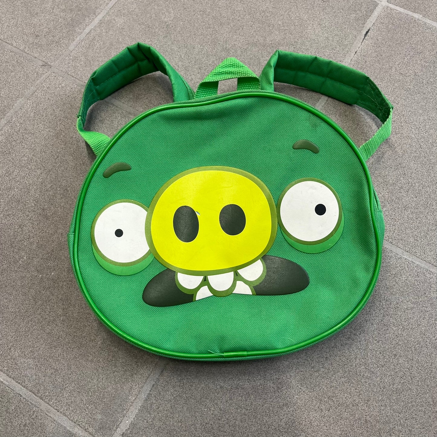 Angry Birds 2010s Bad Piggies Green Backpack is a small circular backpack shaped like the head of a Bad Piggies character. The front features the pig’s scared facial expression, while the side has an Angry Birds logo. The interior includes an all-over pig head monogram lining and an official Angry Birds licensed tag. The backpack is finished with really short adjustable shoulder straps.
