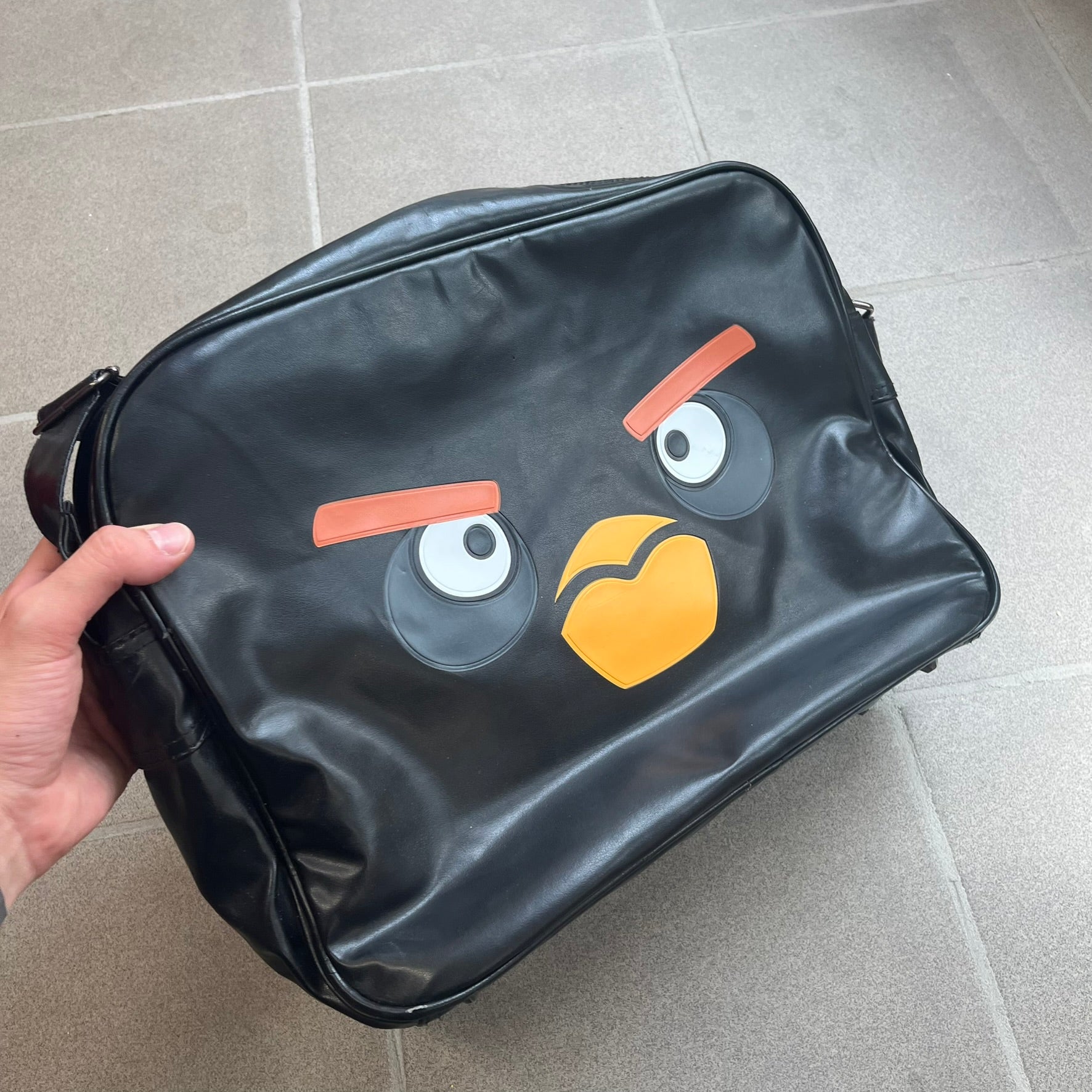 Angry Birds 2010s Black Bomb Bird Shoulder Bag is a black messenger-style bag made from a leather-like material. It features one main compartment with a smaller internal pocket. The front is designed with a large graphic of Bomb, the black Angry Birds character. The interior includes official Angry Birds 2009-2013 licensing.