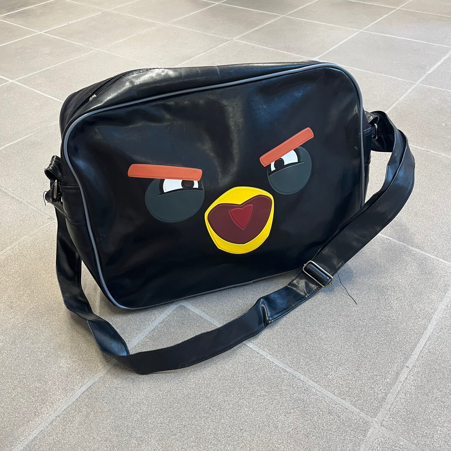 Angry Birds 2010s Black Bomb Bird Shoulder Bag is a black messenger-style bag made from a leather-like material. It features one main compartment with a smaller internal pocket. The front is designed with a large graphic of Bomb, the black Angry Birds character, shown with a screaming expression. The interior includes official Angry Birds 2009-2013 licensing.