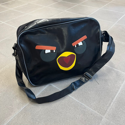 Angry Birds 2010s Black Bomb Bird Shoulder Bag is a black messenger-style bag made from a leather-like material. It features one main compartment with a smaller internal pocket. The front is designed with a large graphic of Bomb, the black Angry Birds character, shown with a screaming expression. The interior includes official Angry Birds 2009-2013 licensing.