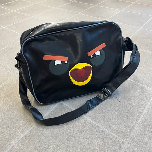 Angry Birds 2010s Black Bomb Bird Shoulder Bag is a black messenger-style bag made from a leather-like material. It features one main compartment with a smaller internal pocket. The front is designed with a large graphic of Bomb, the black Angry Birds character, shown with a screaming expression. The interior includes official Angry Birds 2009-2013 licensing.