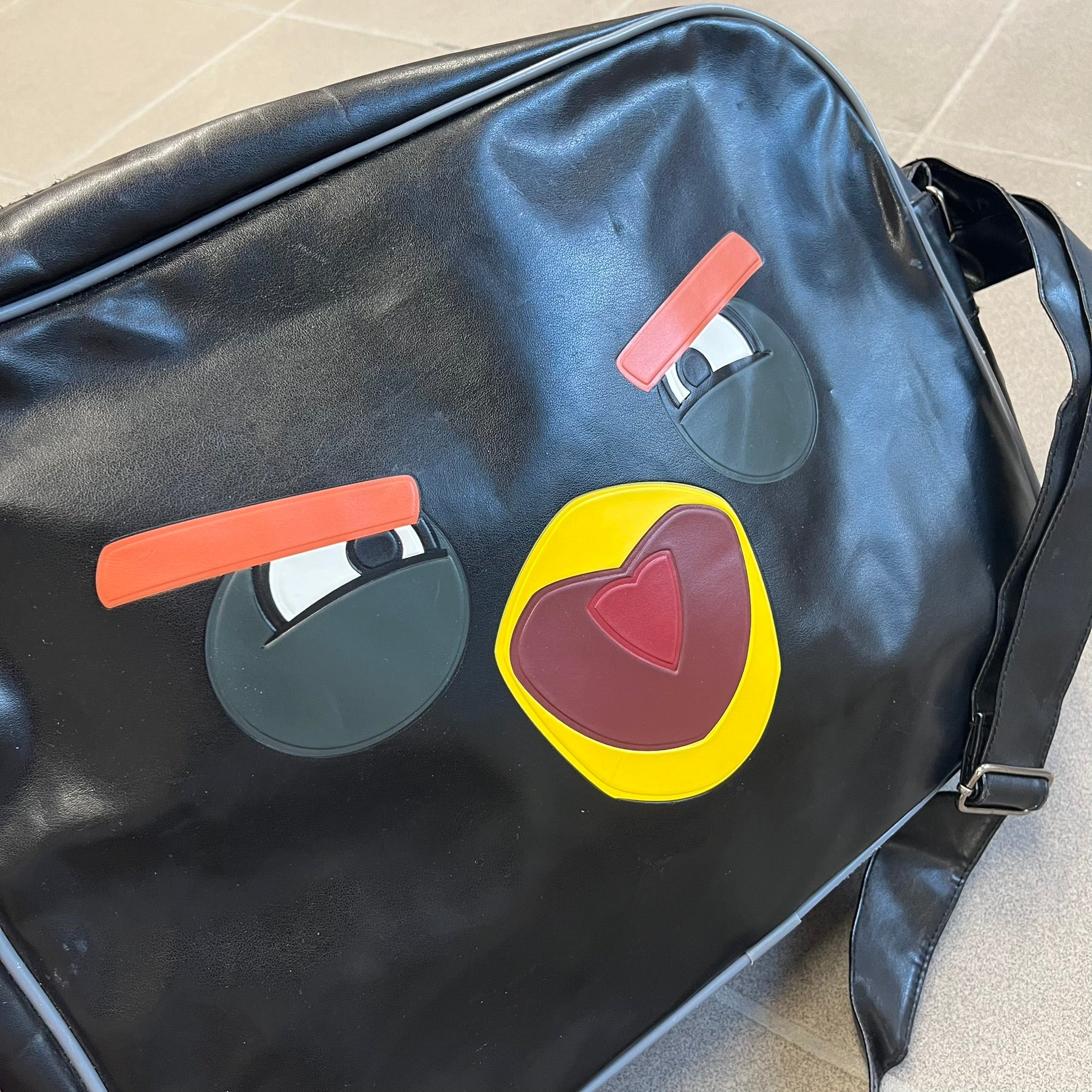 Angry Birds 2010s Black Bomb Bird Shoulder Bag is a black messenger-style bag made from a leather-like material. It features one main compartment with a smaller internal pocket. The front is designed with a large graphic of Bomb, the black Angry Birds character, shown with a screaming expression. The interior includes official Angry Birds 2009-2013 licensing.