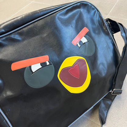 Angry Birds 2010s Black Bomb Bird Shoulder Bag is a black messenger-style bag made from a leather-like material. It features one main compartment with a smaller internal pocket. The front is designed with a large graphic of Bomb, the black Angry Birds character, shown with a screaming expression. The interior includes official Angry Birds 2009-2013 licensing.